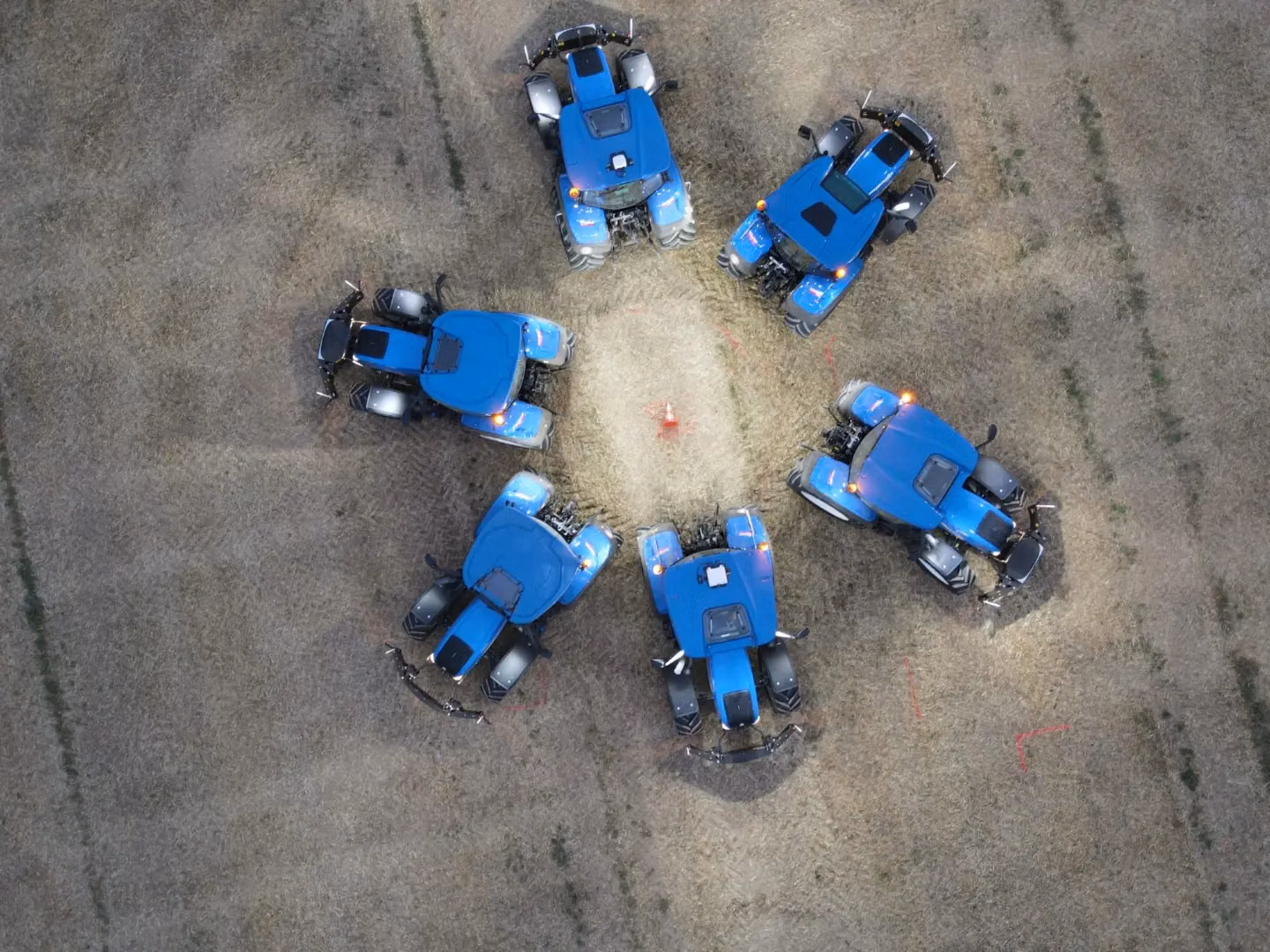Six blue tractors with front weight bumpers arranged in a circle on a dry grassy field seen from above
