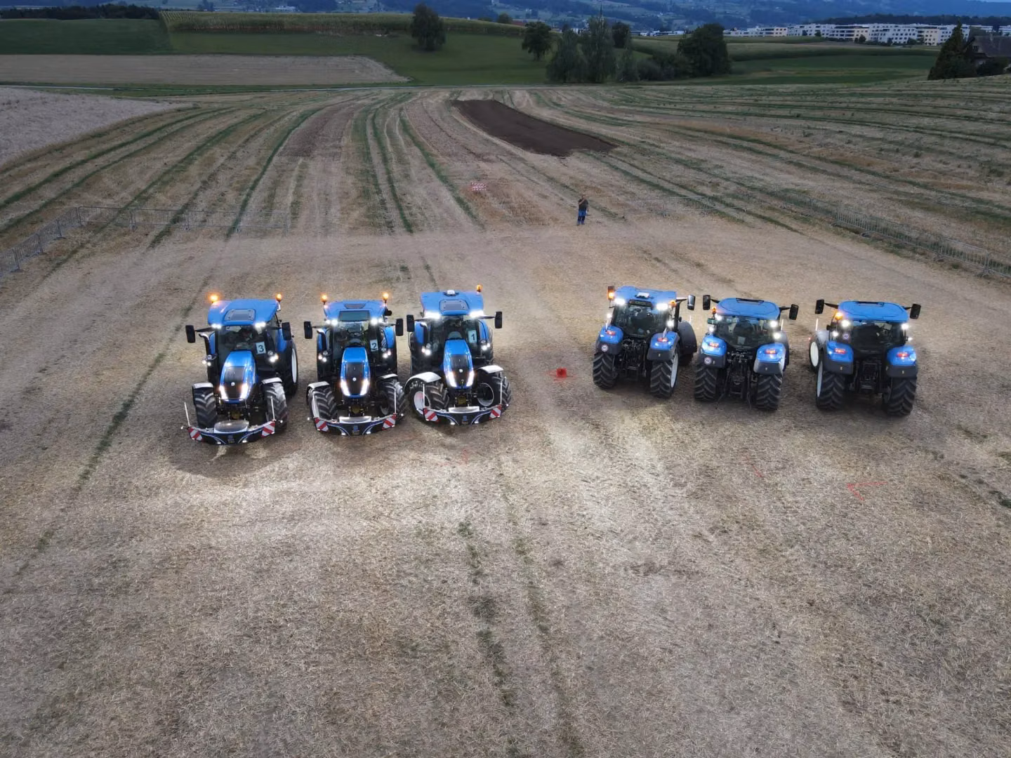 Six blue tractors with illuminated lights are parked in a field, four equipped with front weight bumpers