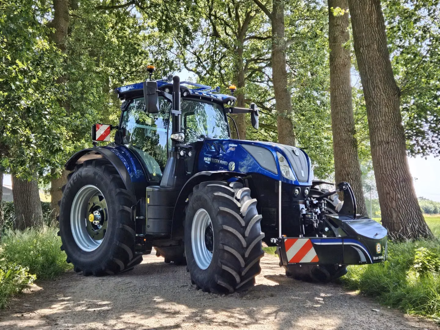 Blue tractor with large tires and a black front weight bumper featuring red and white reflective markings in a wooded area