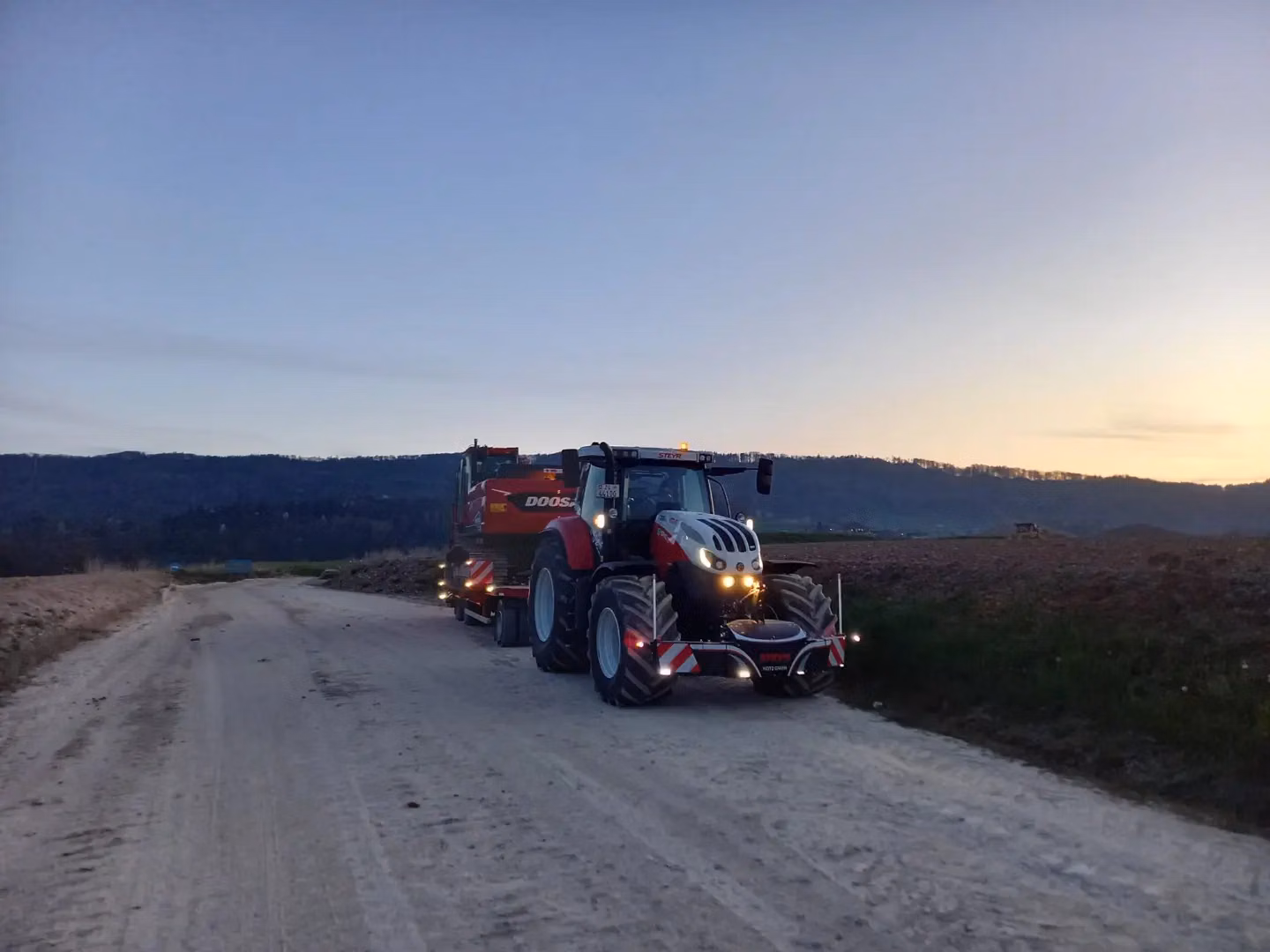 A tractor with headlights on is towing equipment on a dirt road at dusk, fitted with a front bumper marked Steyr.