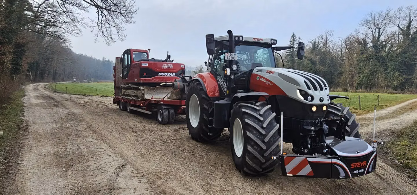 A Steyr tractor with a front weight bumper attached is towing a trailer carrying a red excavator on a dirt road