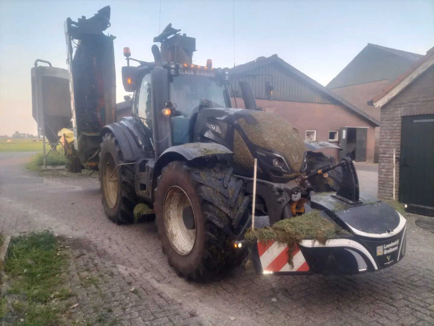 A tractor with grass-covered tires and a front weight bumper is parked on a farmyard near agricultural buildings