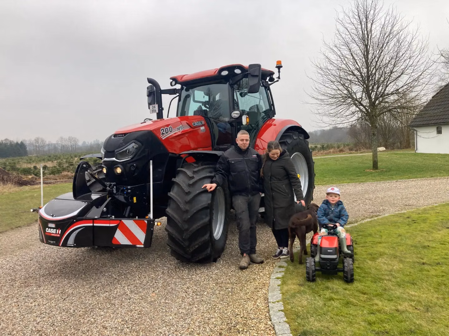A red tractor with a front weight bumper parked on gravel with two adults, a child on a toy tractor, and a dog nearby