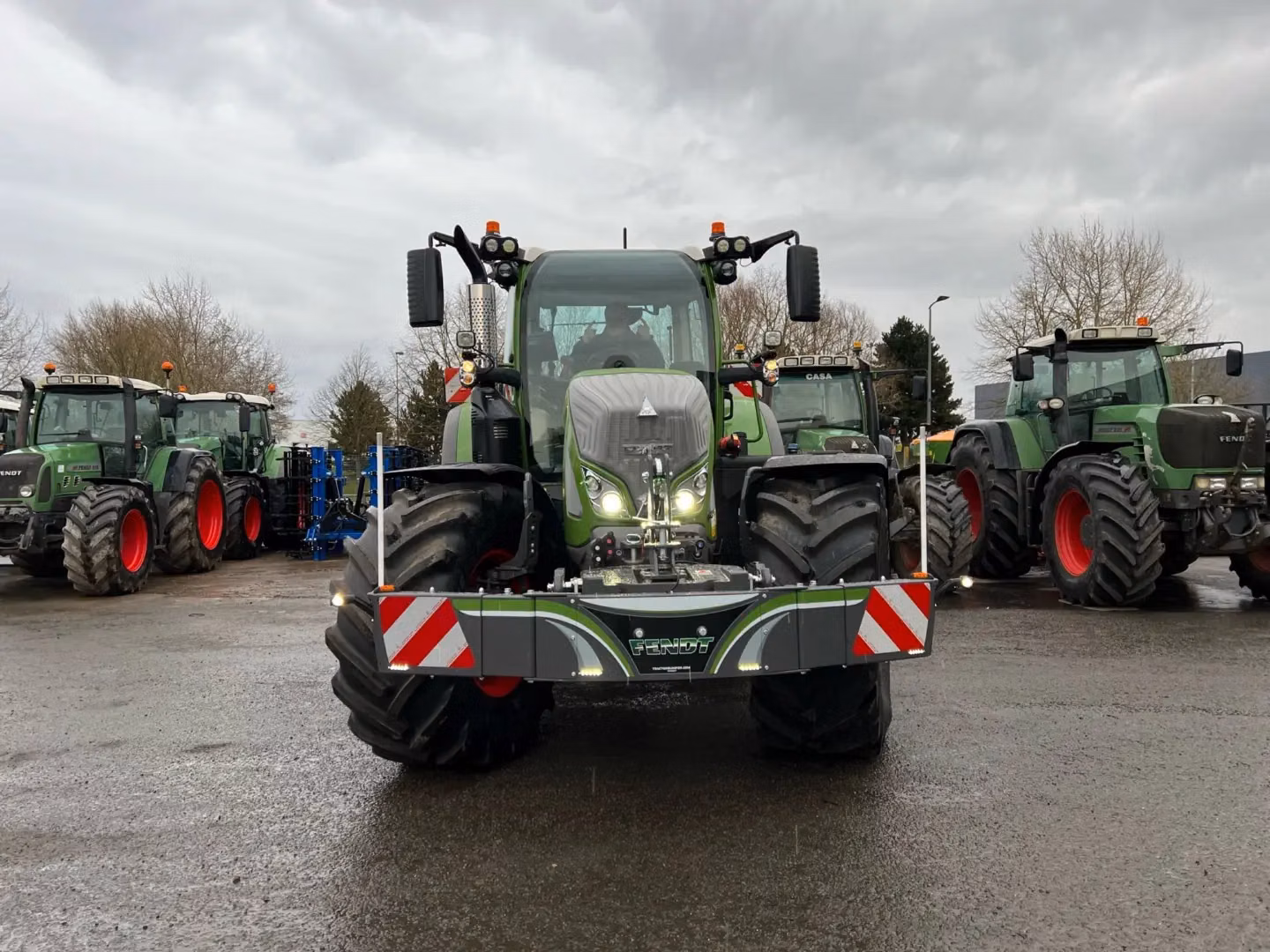 A green tractor with a front weight bumper is parked on wet pavement, with three more tractors in the background