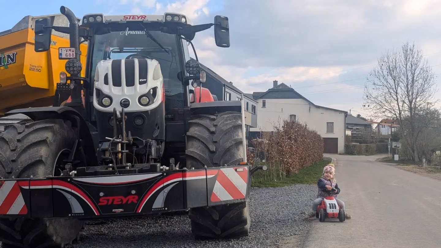 Large tractor with a front weight bumper parked on gravel beside a child riding a small toy tractor on the road