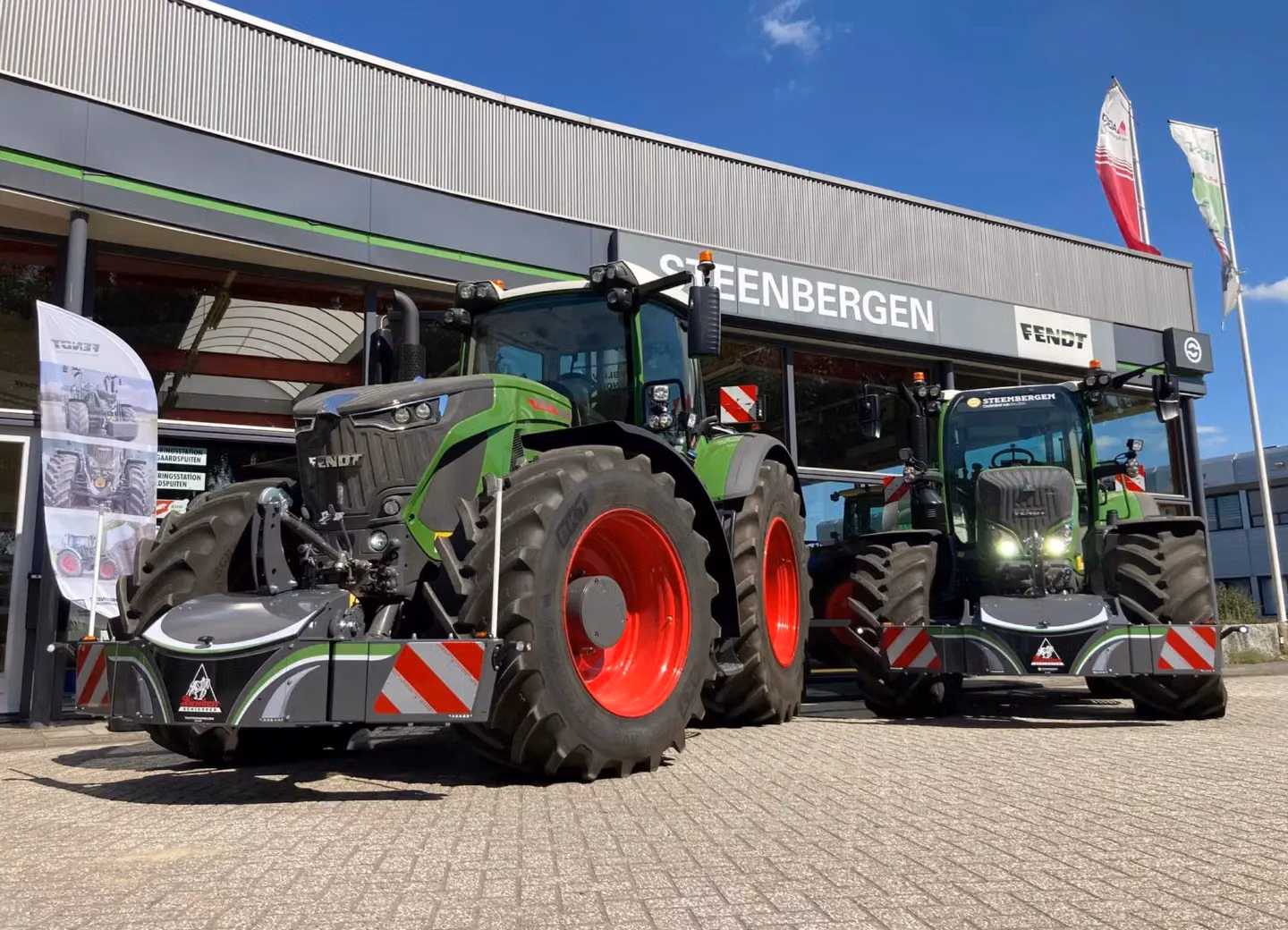 Two green tractors with red wheels fitted with front weight bumpers are parked outside a building with Fendt signs