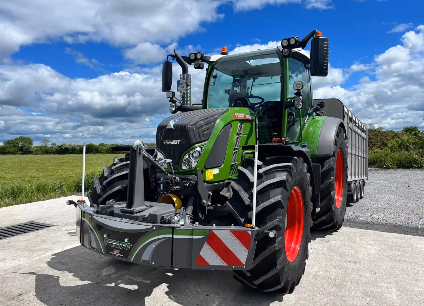 Green tractor with red wheels and a large front weight bumper, parked on concrete beside a grassy field