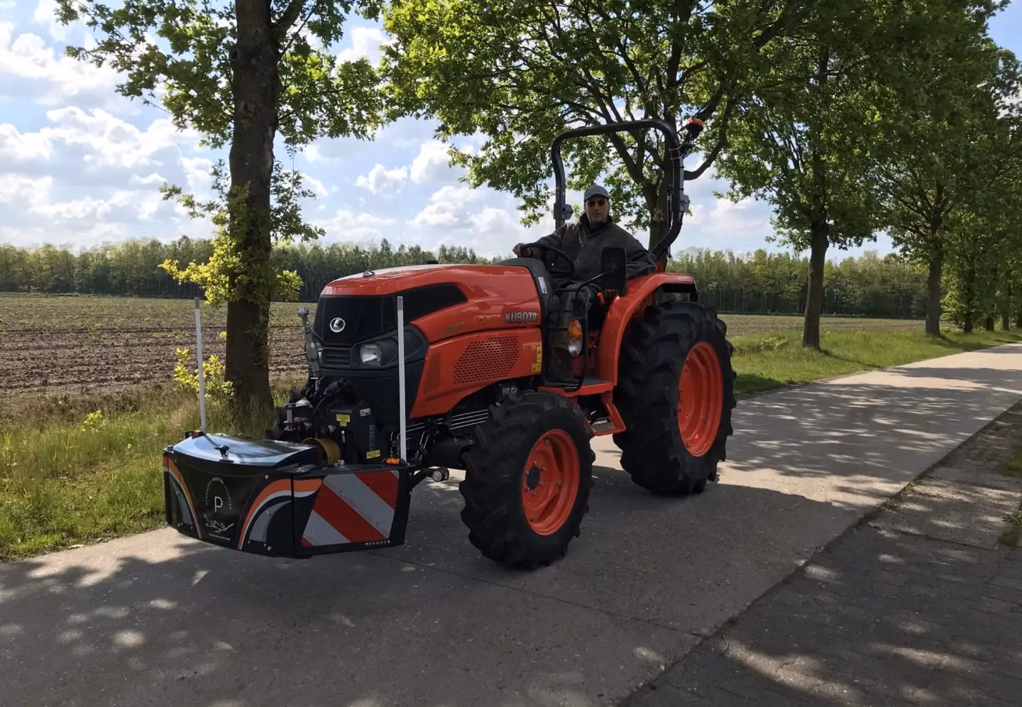 A man drives an orange tractor with a mounted front weight bumper on a paved road beside a field and trees