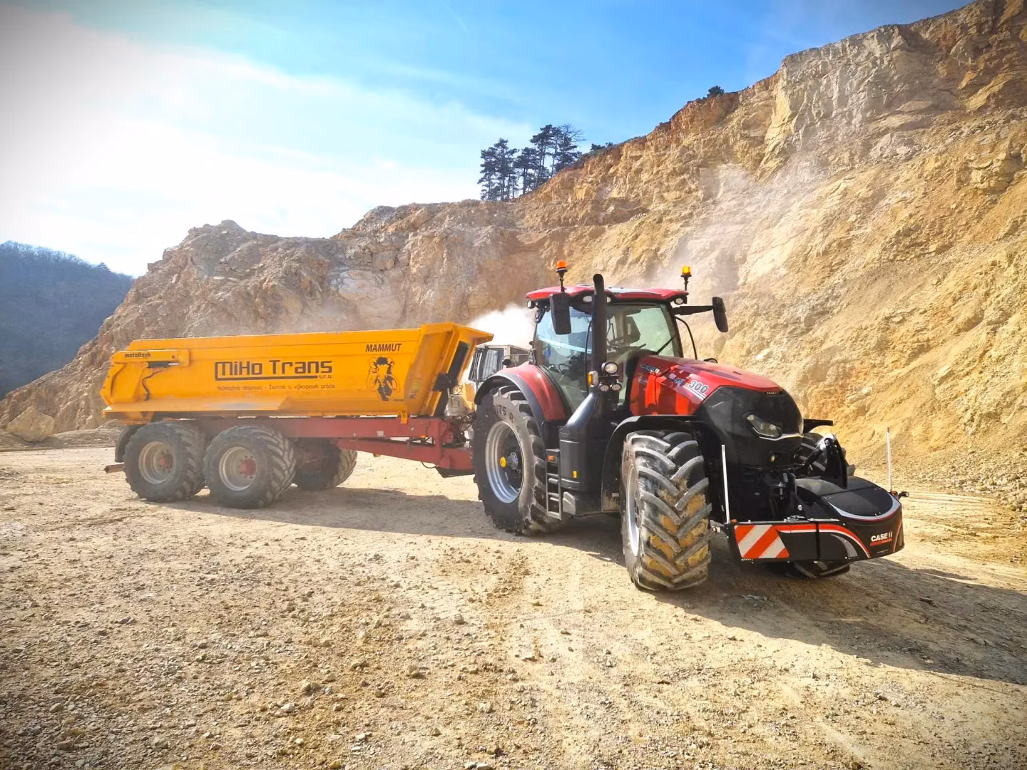 Red tractor with a front weight bumper attached, pulling a large yellow trailer on a dirt quarry site
