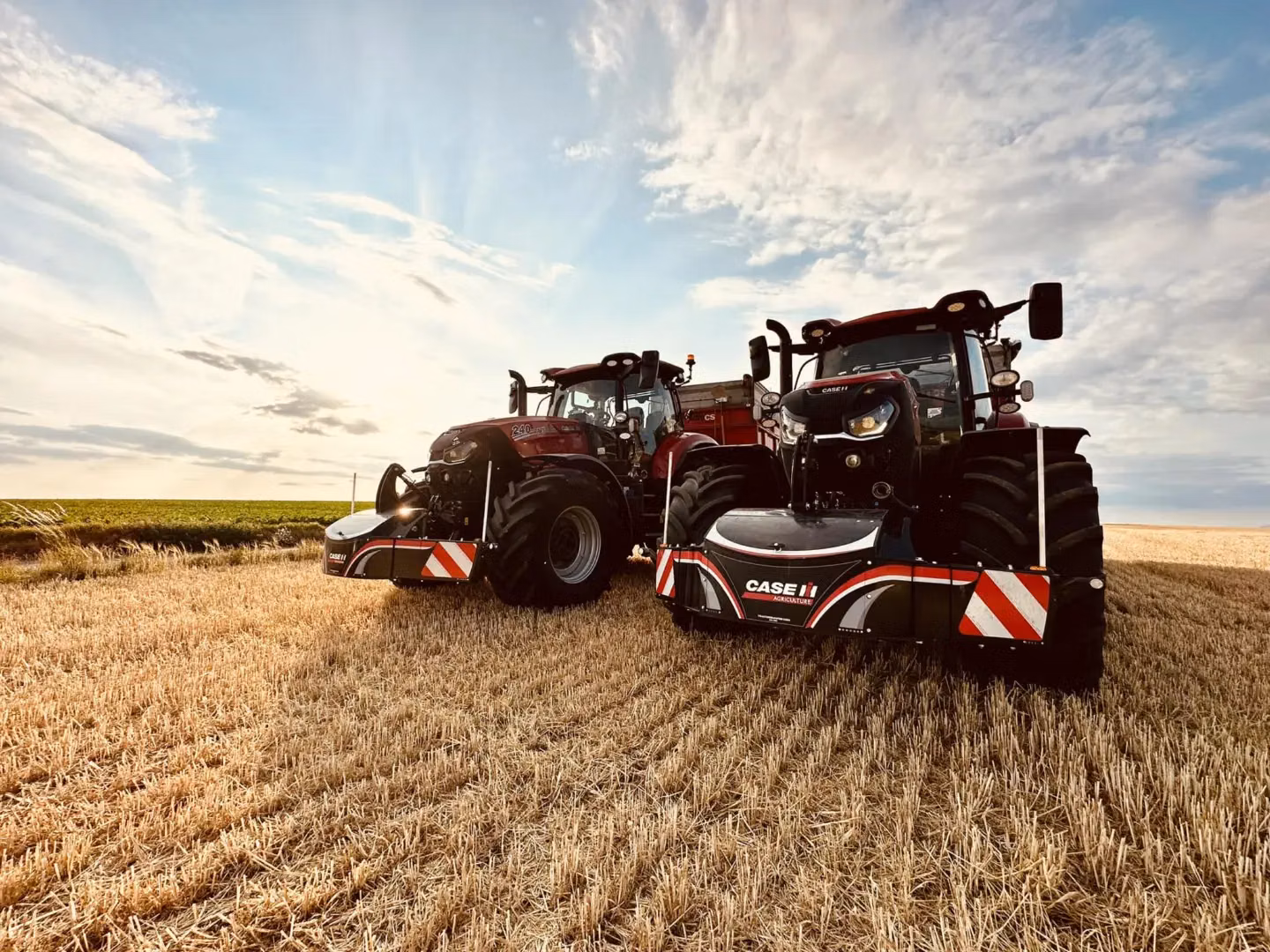 Two red tractors with CASE IH branded front weight bumpers parked on a harvested field under a partly cloudy sky