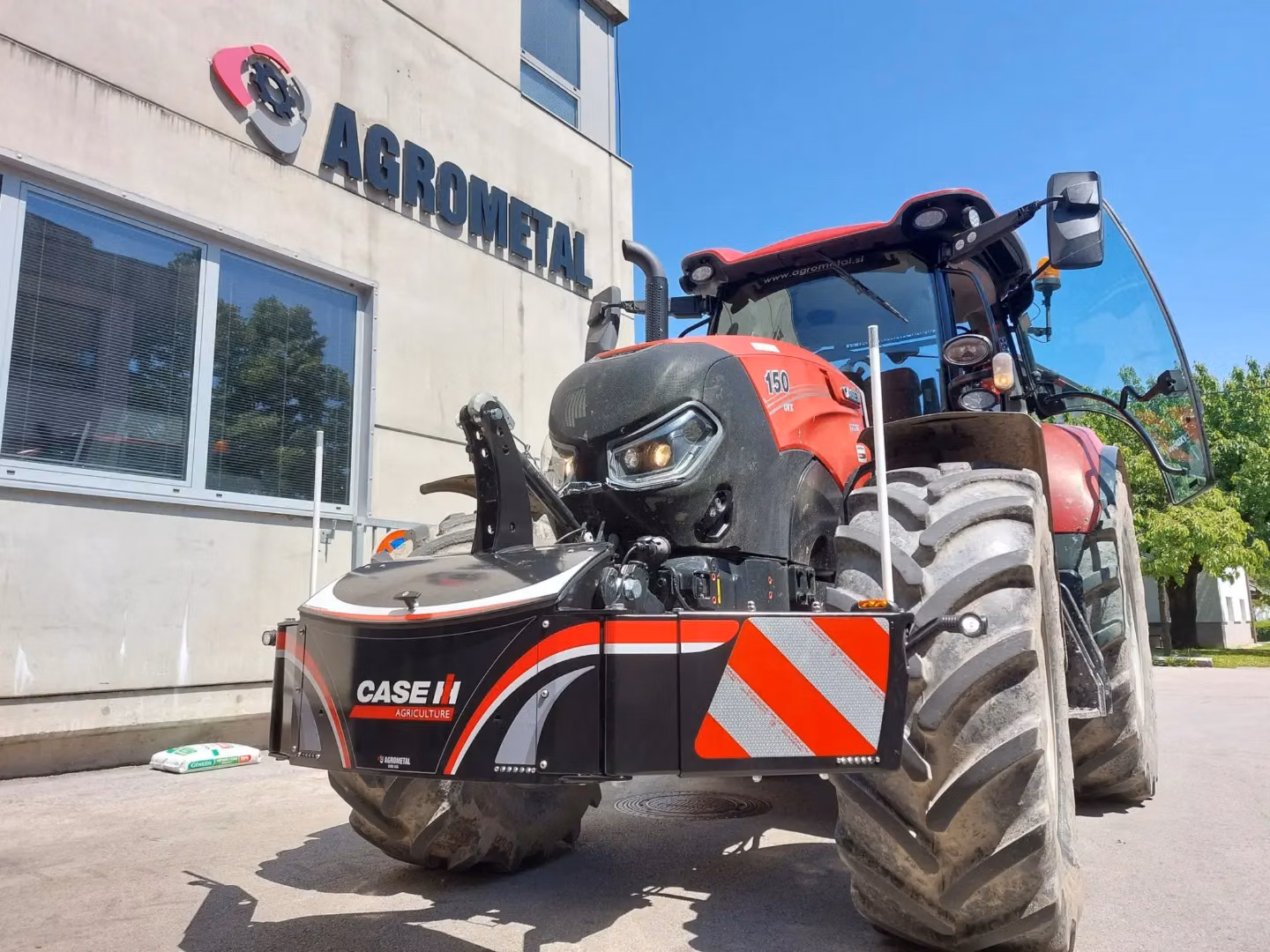Red tractor with a CASE IH branded front weight bumper parked outside a building with AGROMETAL signage