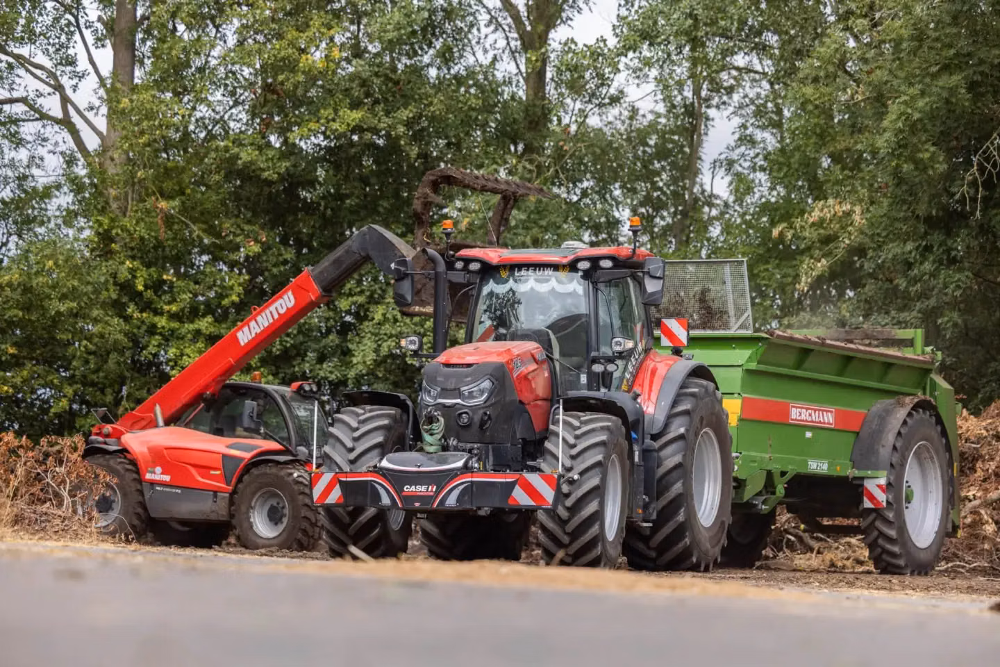 A red tractor with a front weight bumper is parked between a red loader and a green trailer filled with debris