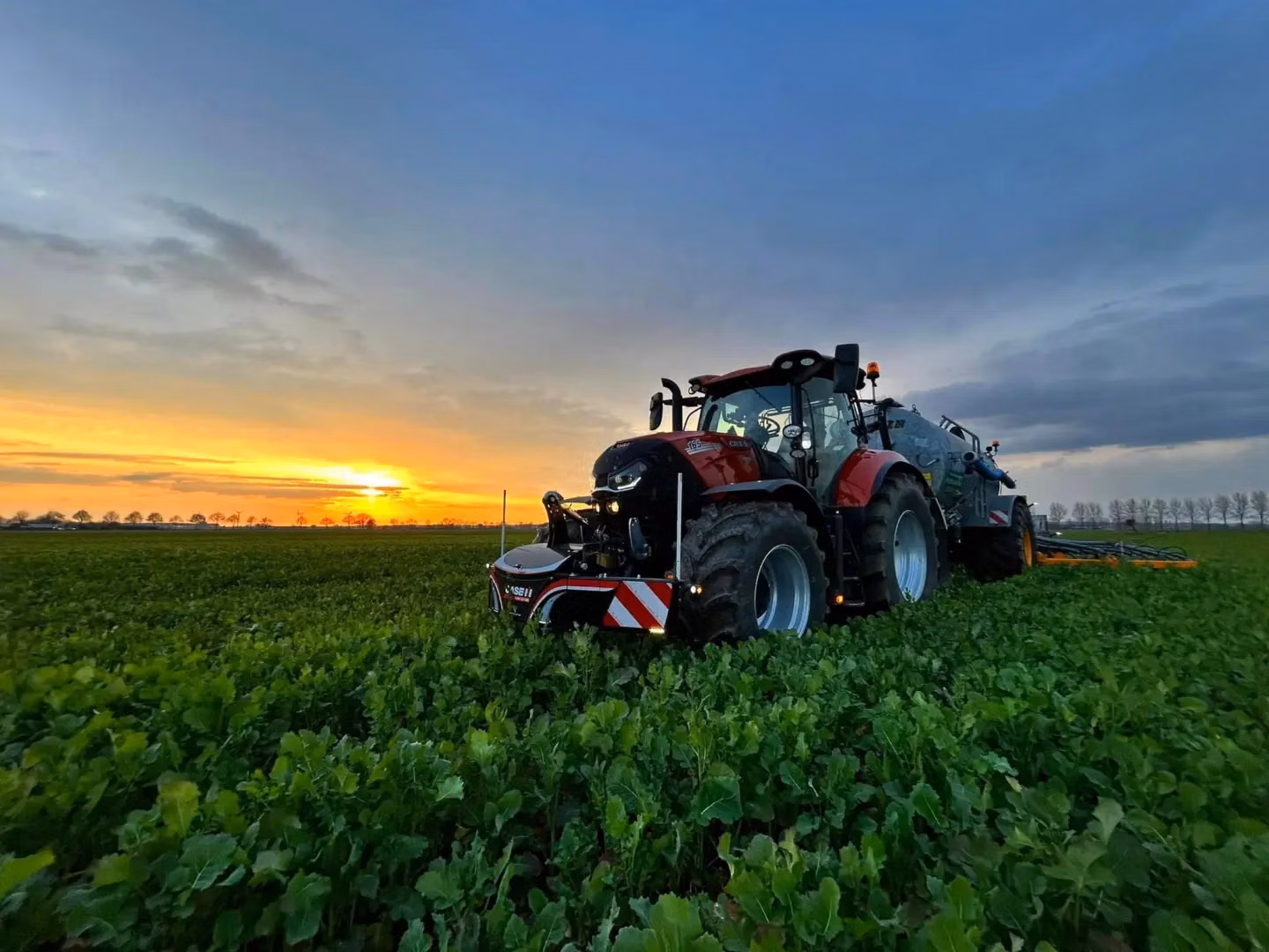 Red tractor with a front weight bumper and large rear attachment driving through a green field at sunset