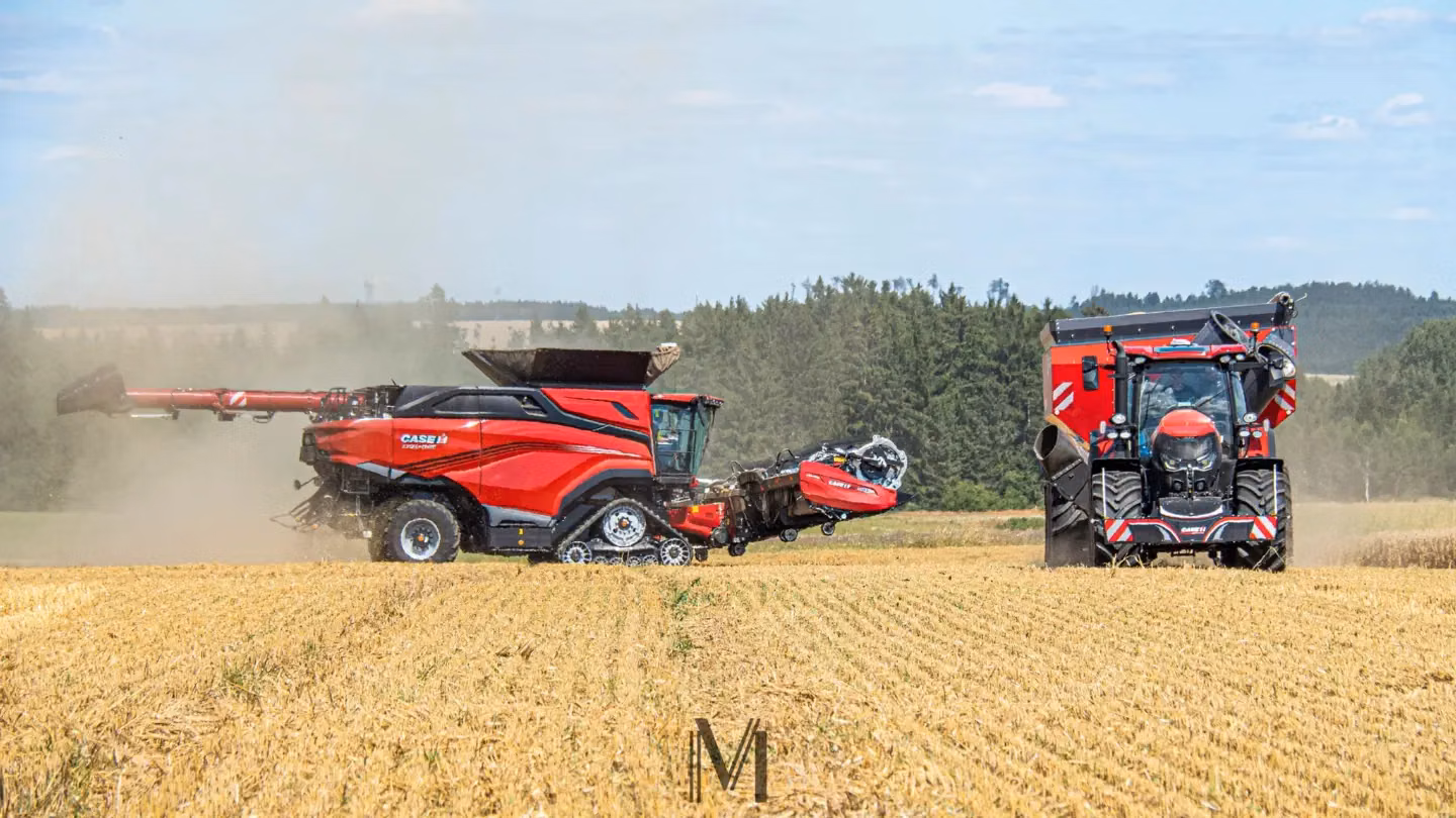 A red combine harvester and a red tractor with a front weight bumper work in a harvested field with trees in the background