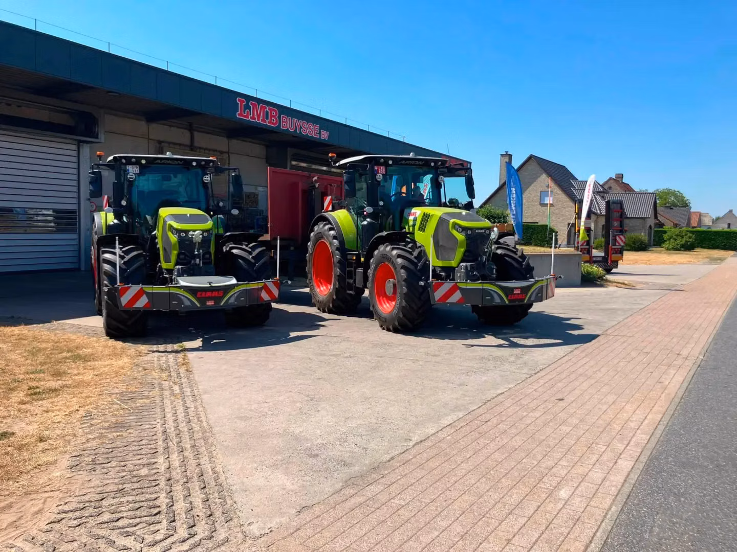 Two green tractors with front weight bumpers are parked on concrete outside a building under a blue sky