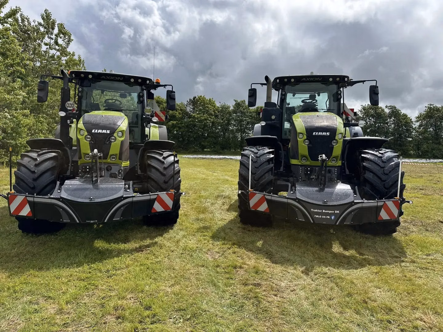 Two green tractors with front weight bumpers are parked side by side on grass with trees and a cloudy sky behind.