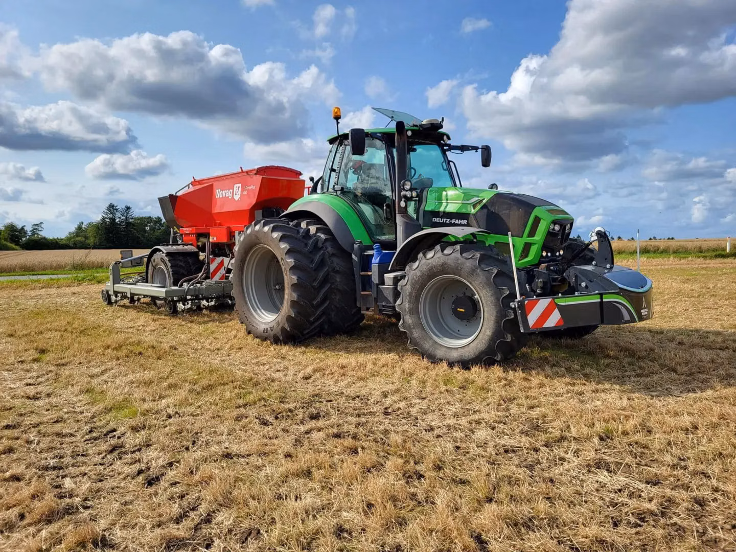 Green tractor with a front weight bumper and large rear implement attached, parked on a harvested field