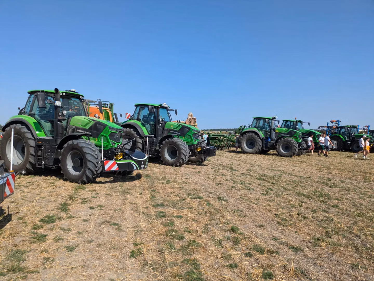 Several green tractors with front weight bumpers are parked on a dry field with people walking nearby