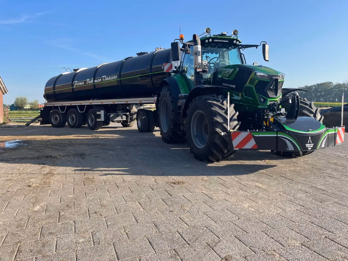 A green tractor with a large front weight bumper is attached to a black tanker trailer on a paved surface