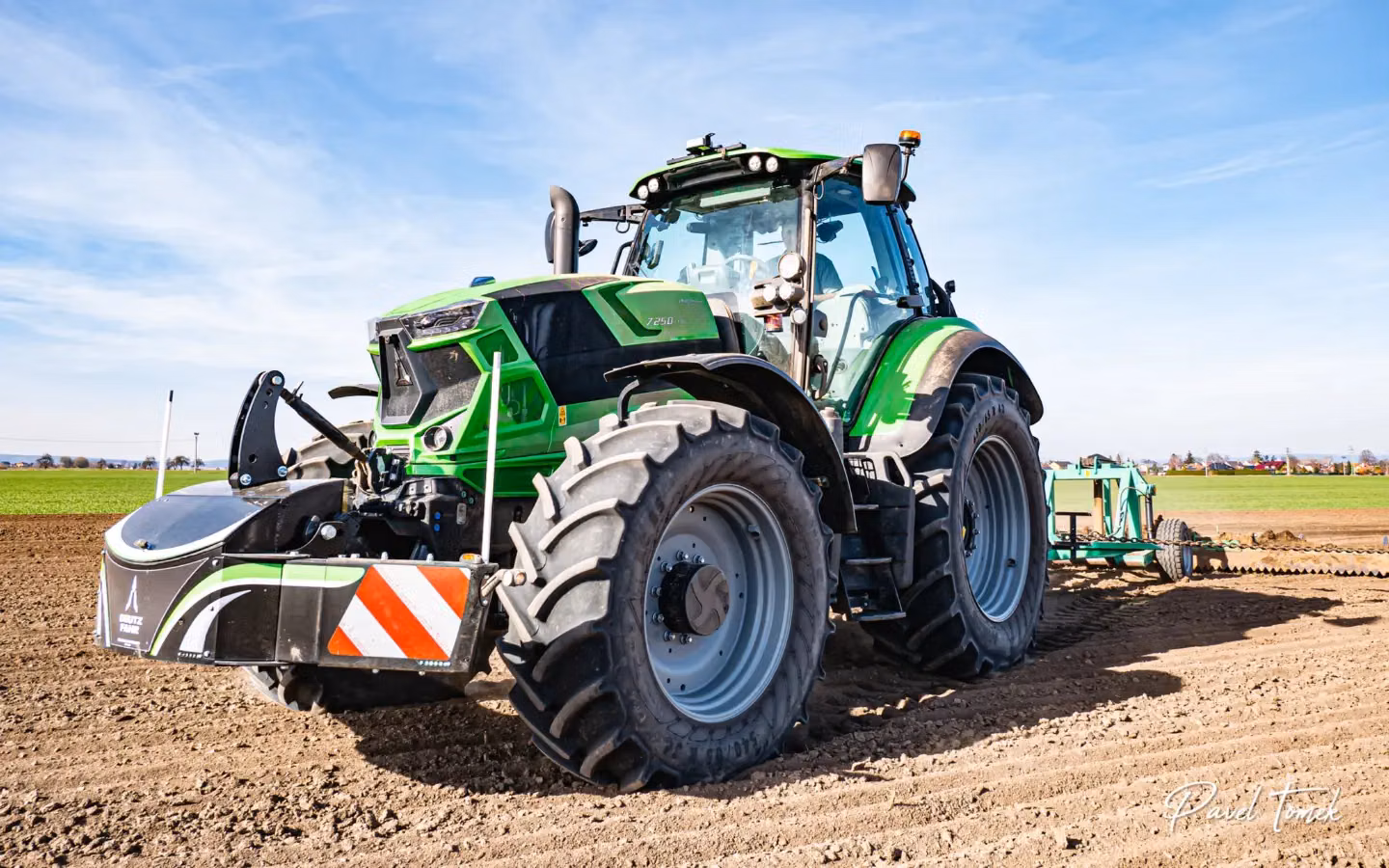 Green tractor with a front weight bumper and warning stripes working on a field under a blue sky