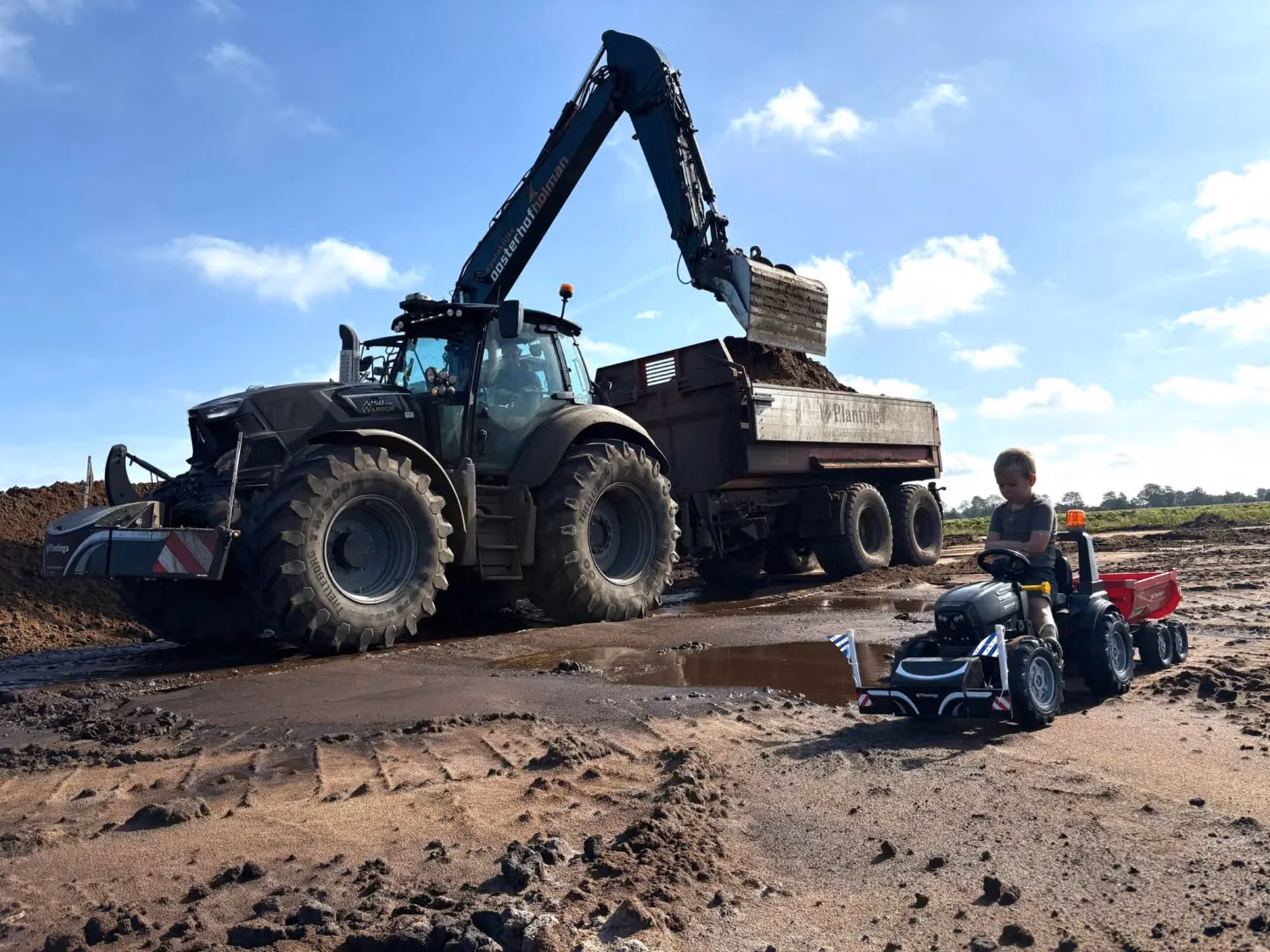 Large tractor with a front weight bumper and excavator loading soil into a trailer, child on toy tractor in foreground