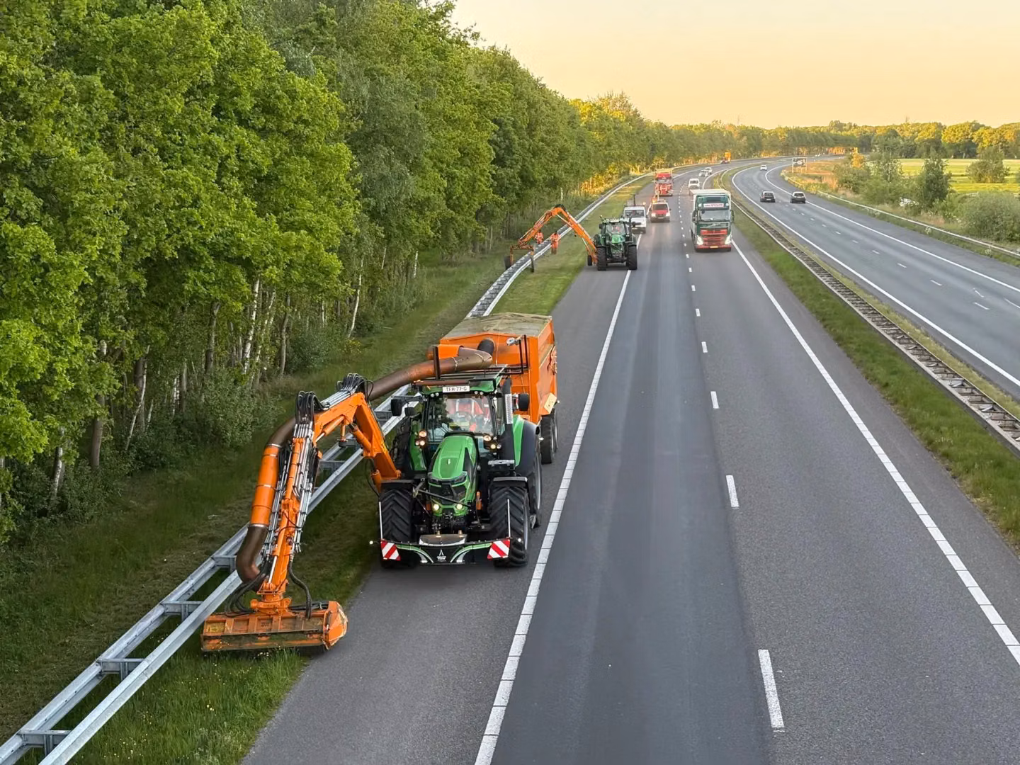 A green tractor with a front weight bumper and mowing attachment trims grass beside a highway with other vehicles nearby