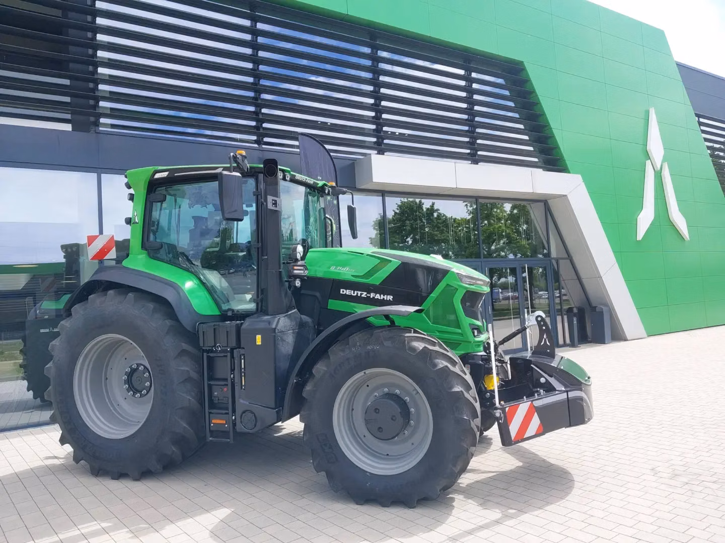 Green tractor with large front weight bumper and red-white safety stripes parked outside a modern green building