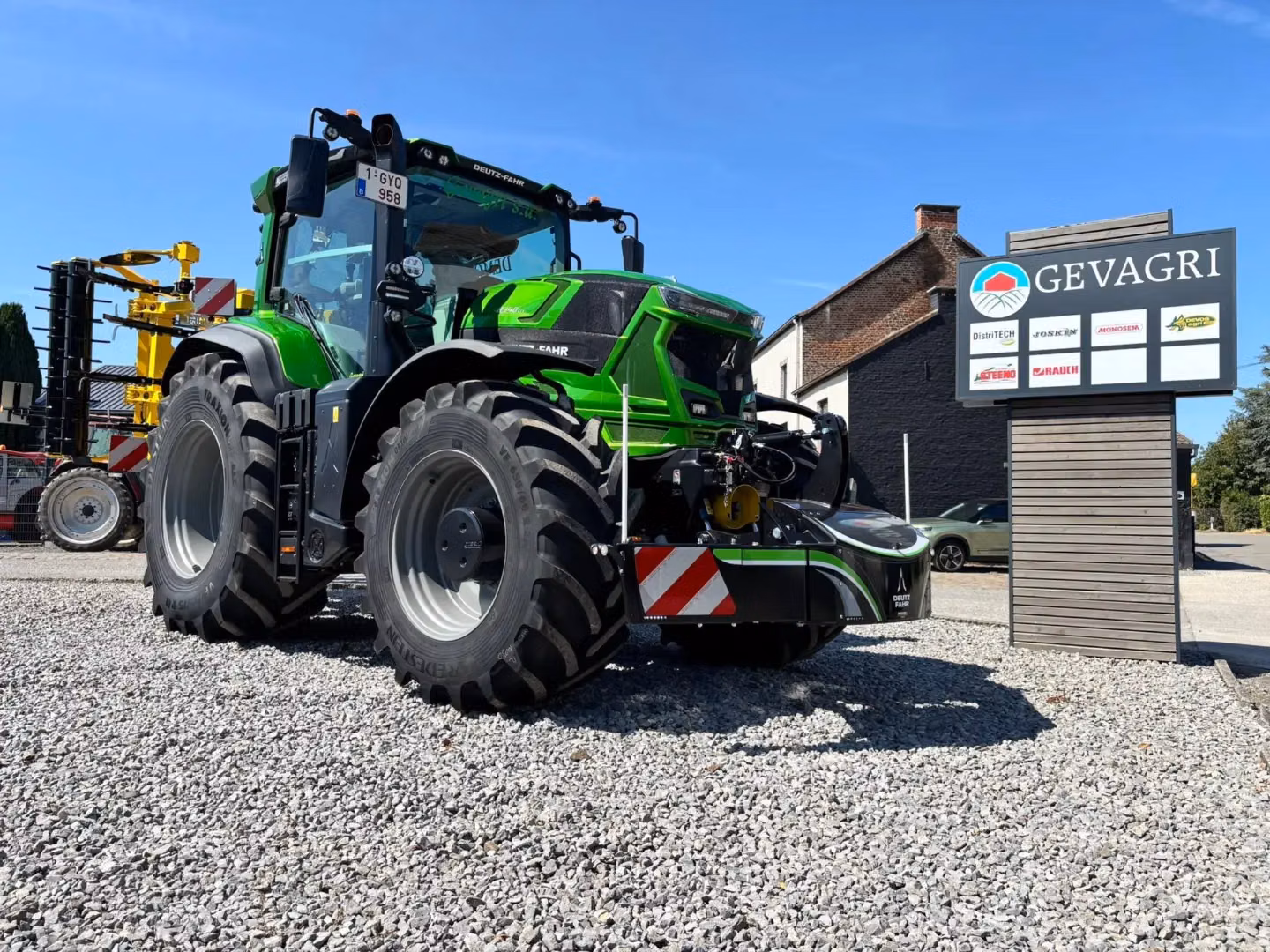 Green tractor with large front weight bumper on gravel beside a sign reading GEVAGRI under a clear blue sky