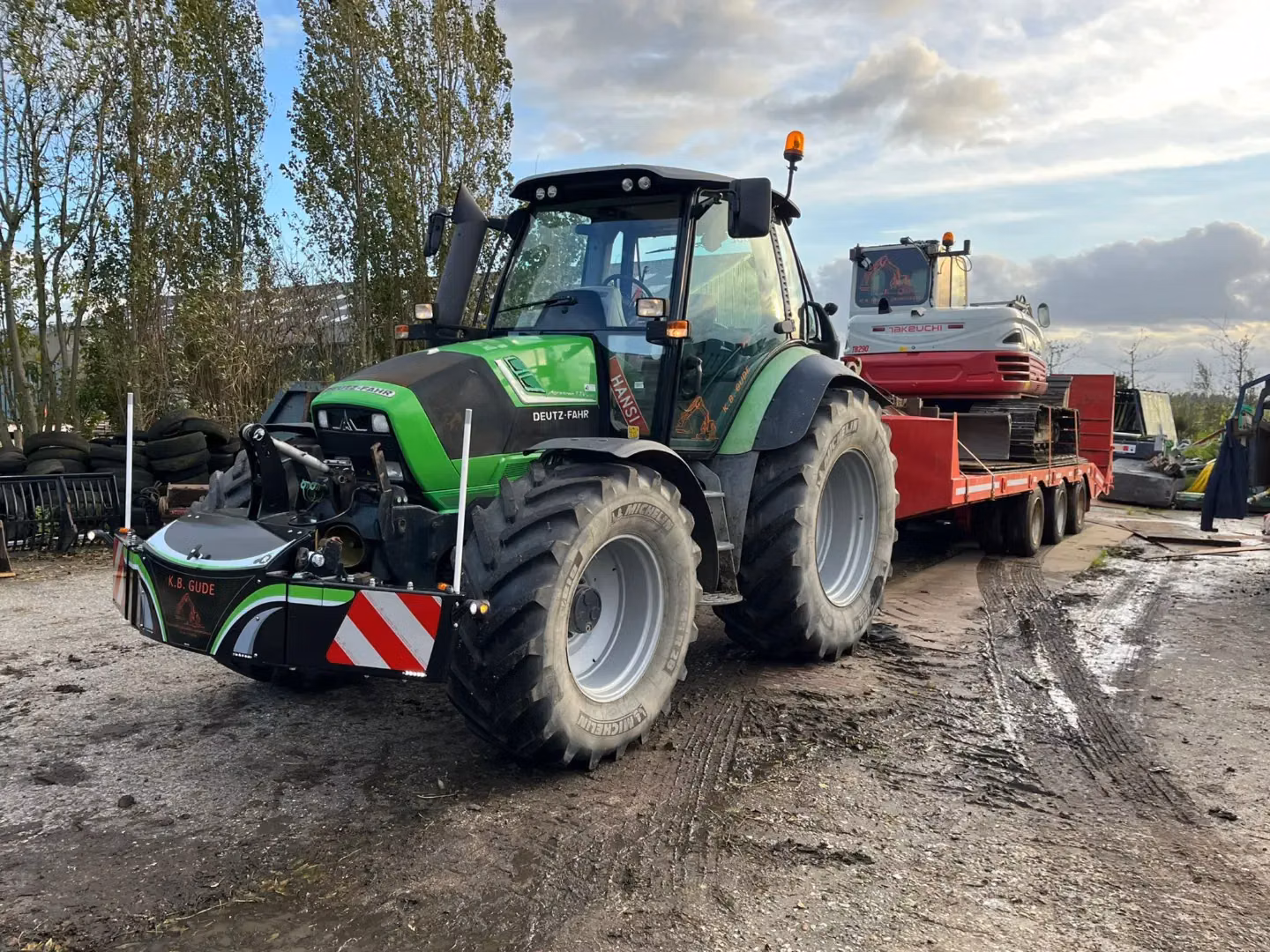 A green tractor with a front weight bumper is parked on a muddy yard, towing a trailer with a red excavator.