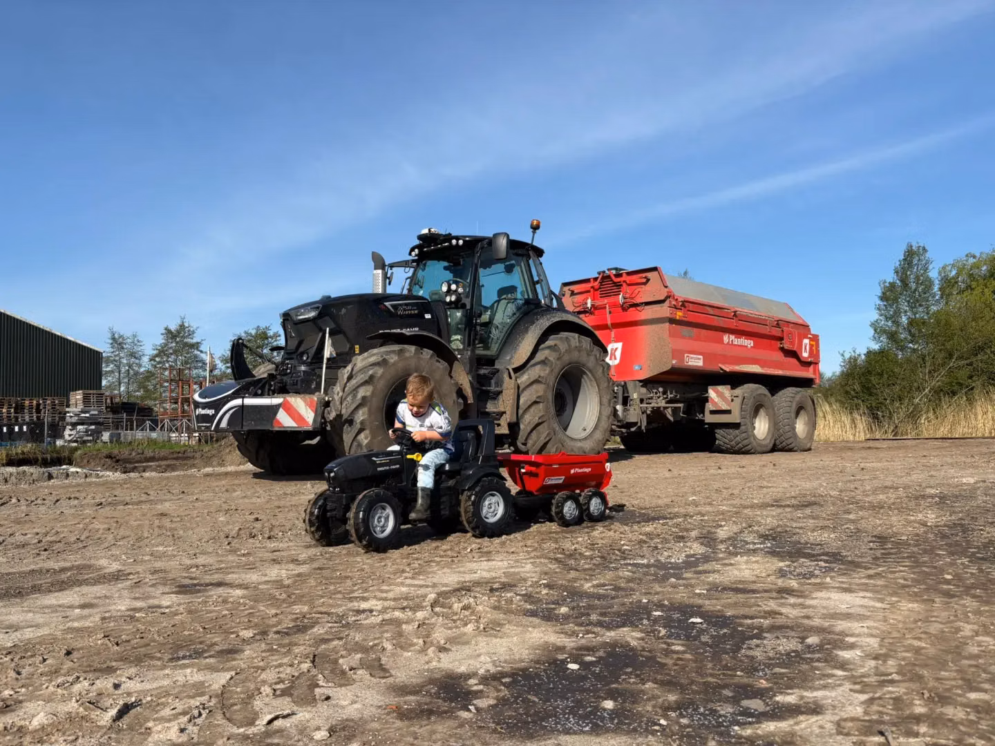 A child sits on a black toy tractor in front of a parked black tractor with a front weight bumper and a red trailer