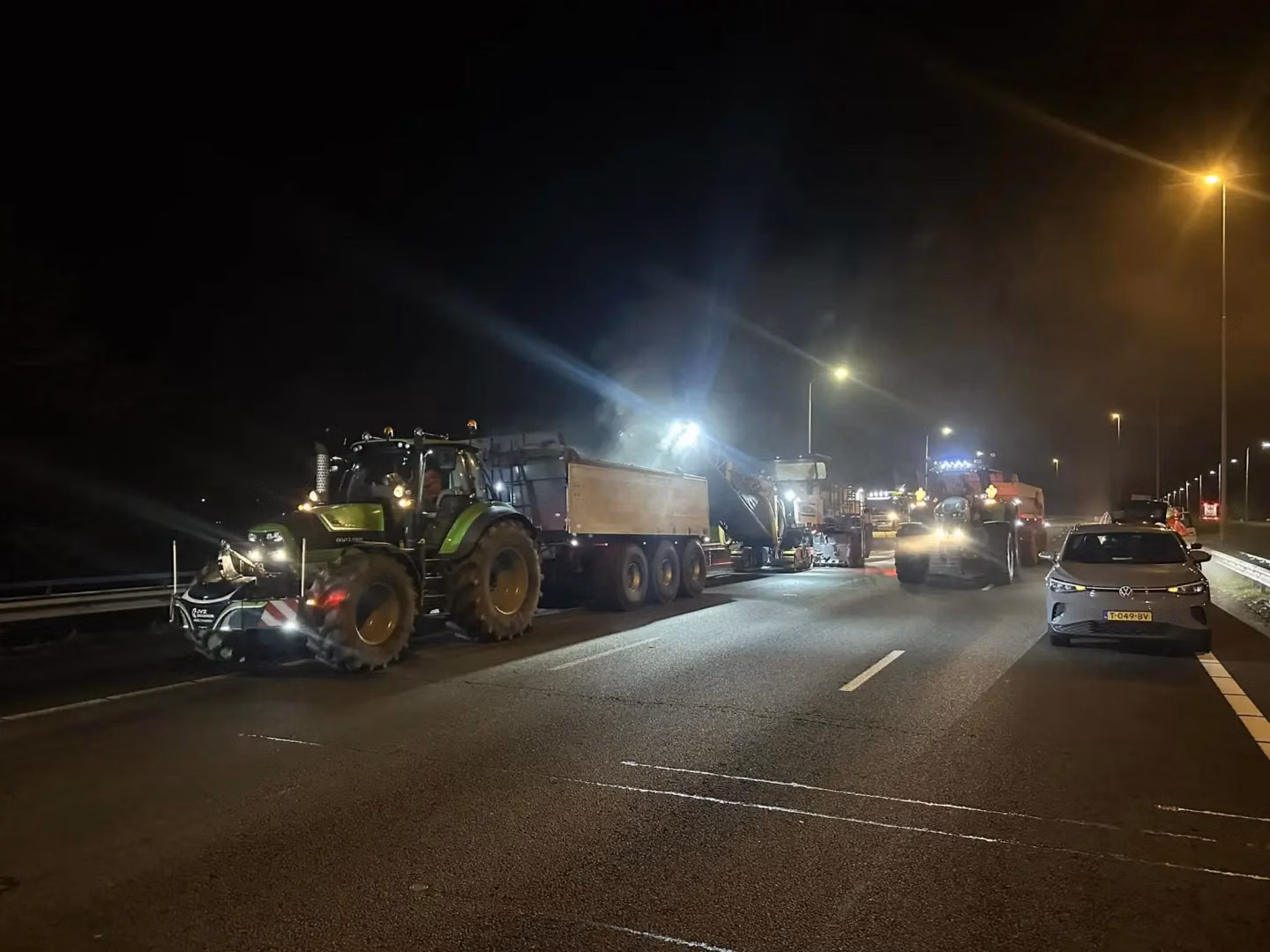 A tractor with a front weight bumper is working at night on a highway with construction vehicles and lights