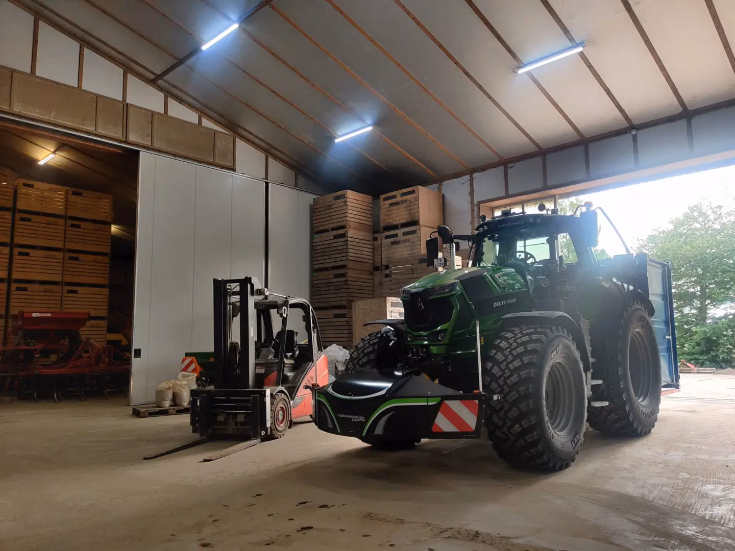 A green tractor with a front weight bumper and a forklift are parked inside a warehouse with stacked crates.