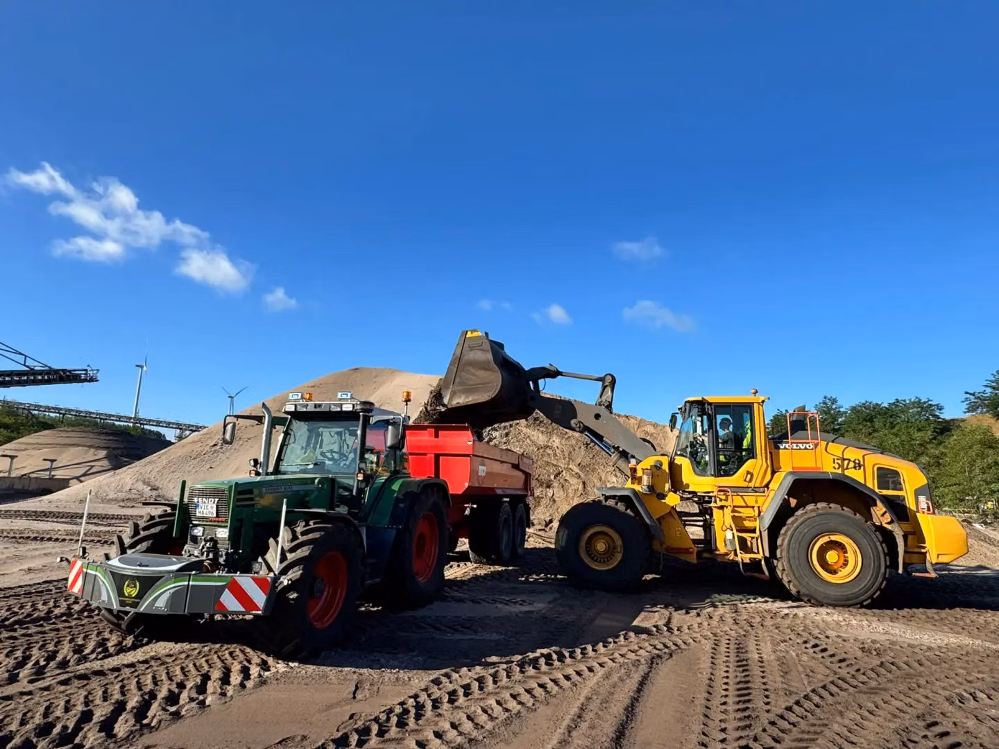 A green tractor with a front weight bumper and red trailer is parked beside a yellow wheel loader at a sandy worksite