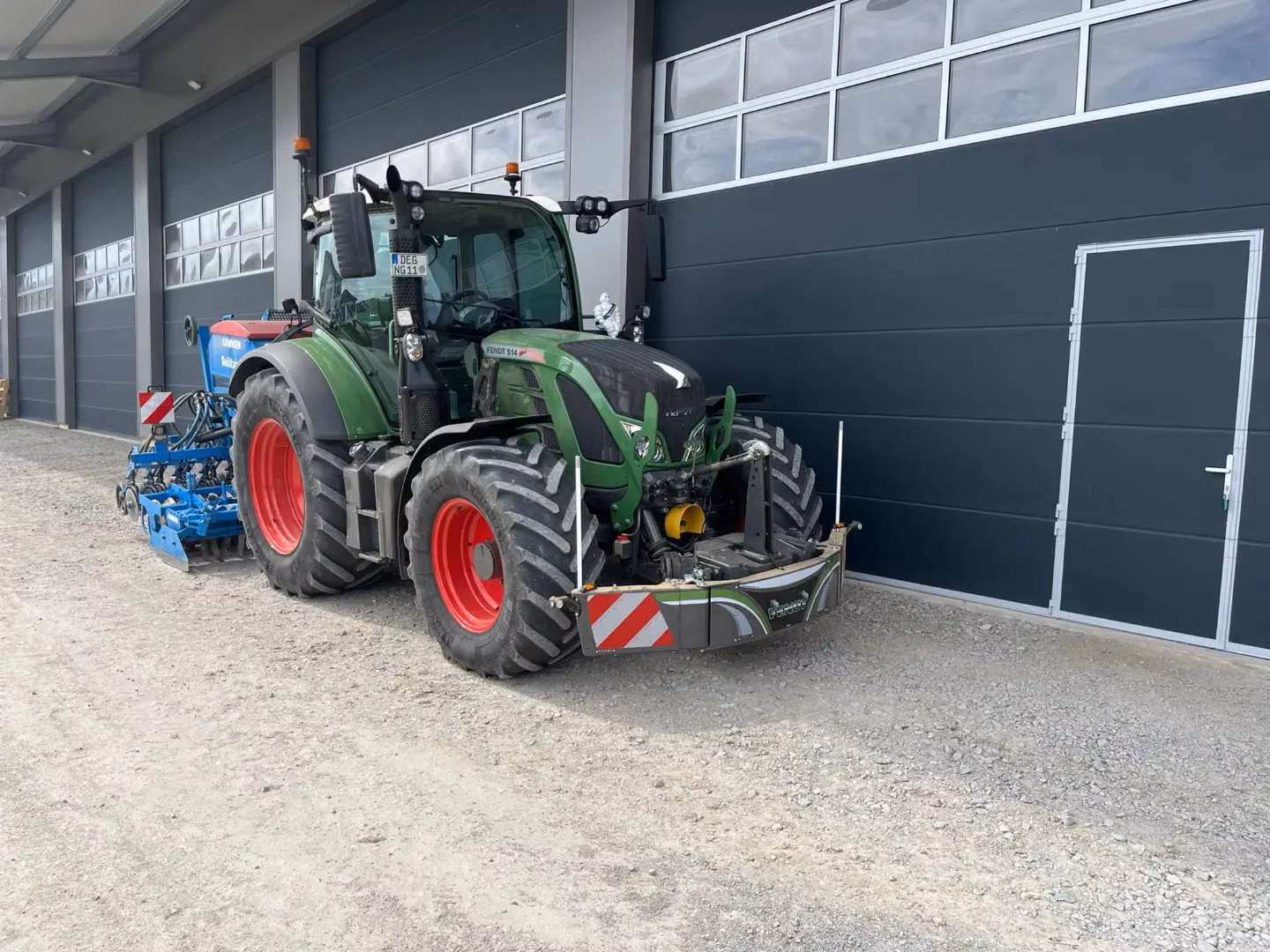 Green tractor with red wheels fitted with a front weight bumper, parked outside a building with grey doors