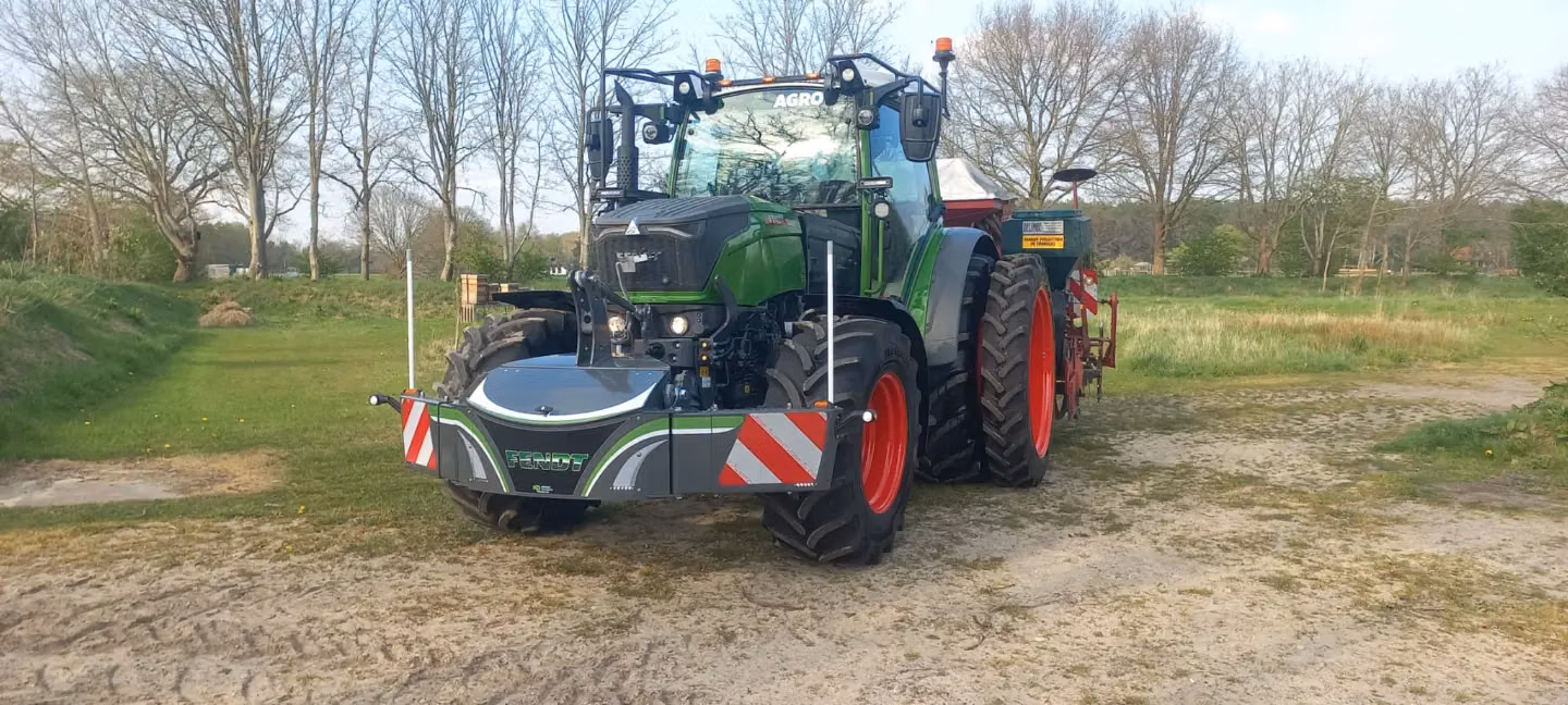Green tractor with red wheels equipped with a front weight bumper on a grassy area with trees in the background