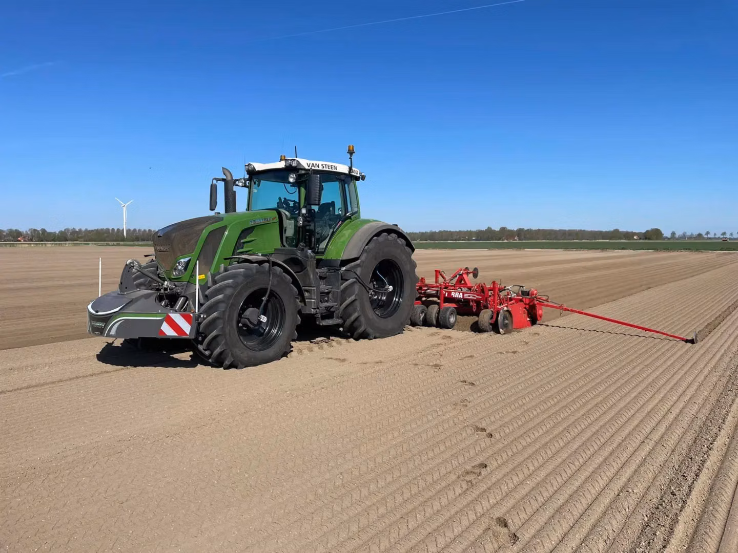 Green tractor with a front weight bumper working in a plowed field, pulling red agricultural equipment