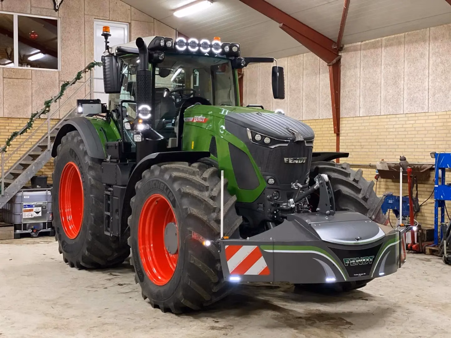 A green tractor with red wheels inside a workshop, fitted with a Fendt front weight bumper displaying reflective stripes