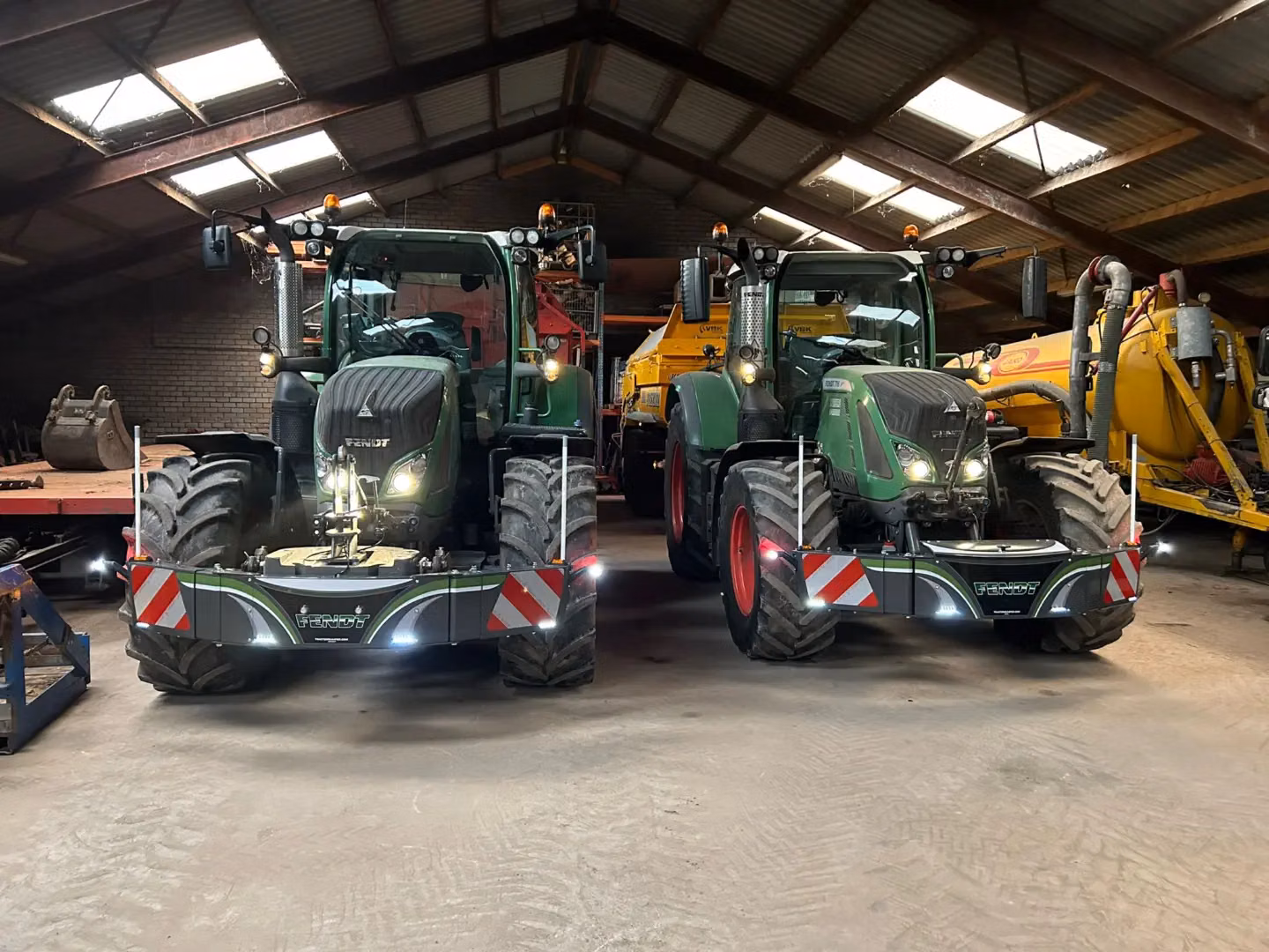 Two green tractors fitted with front weight bumpers featuring reflective red and white hazard stripes inside a shed