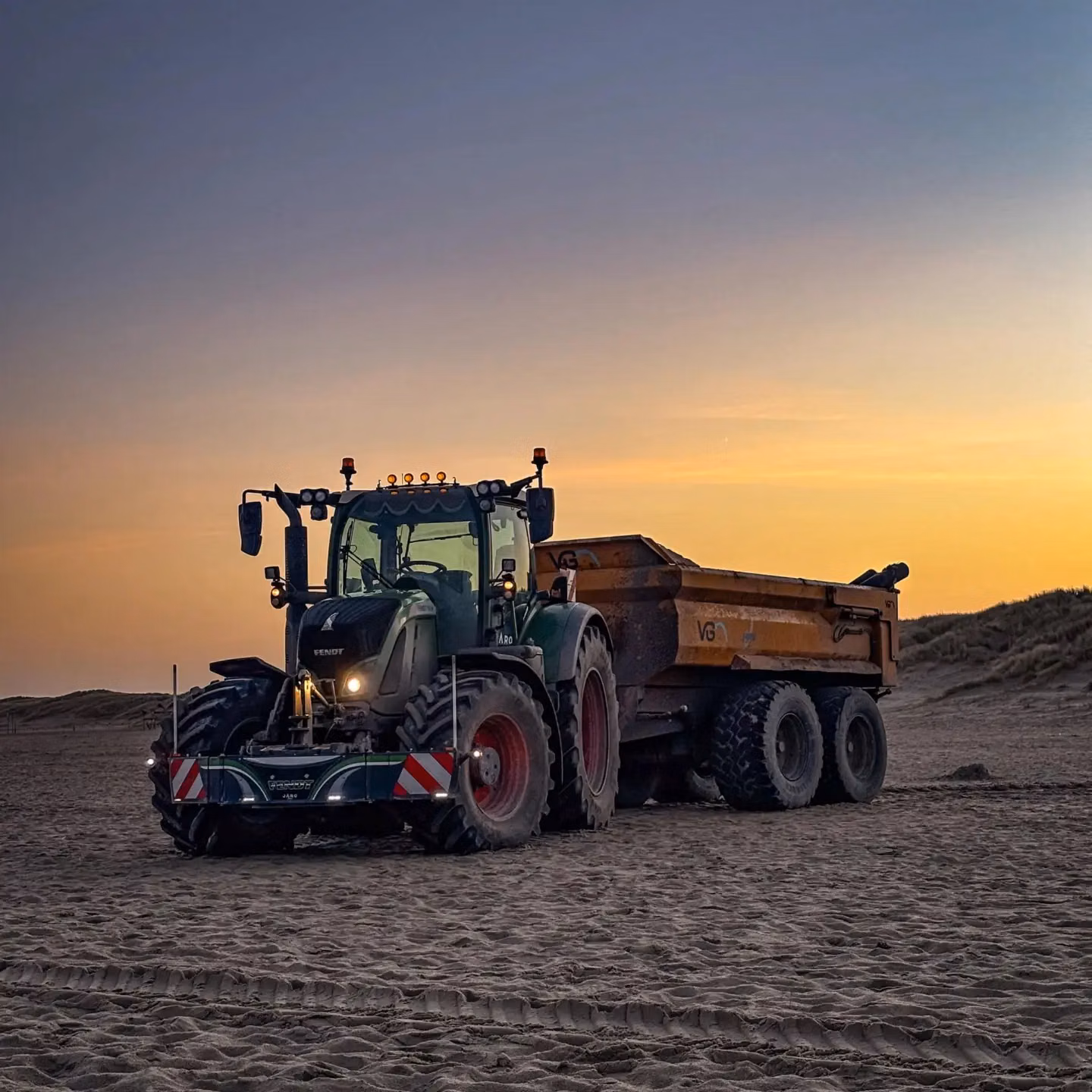 Tractor with a front weight bumper and lights attached, pulling a large trailer on sandy terrain at sunset