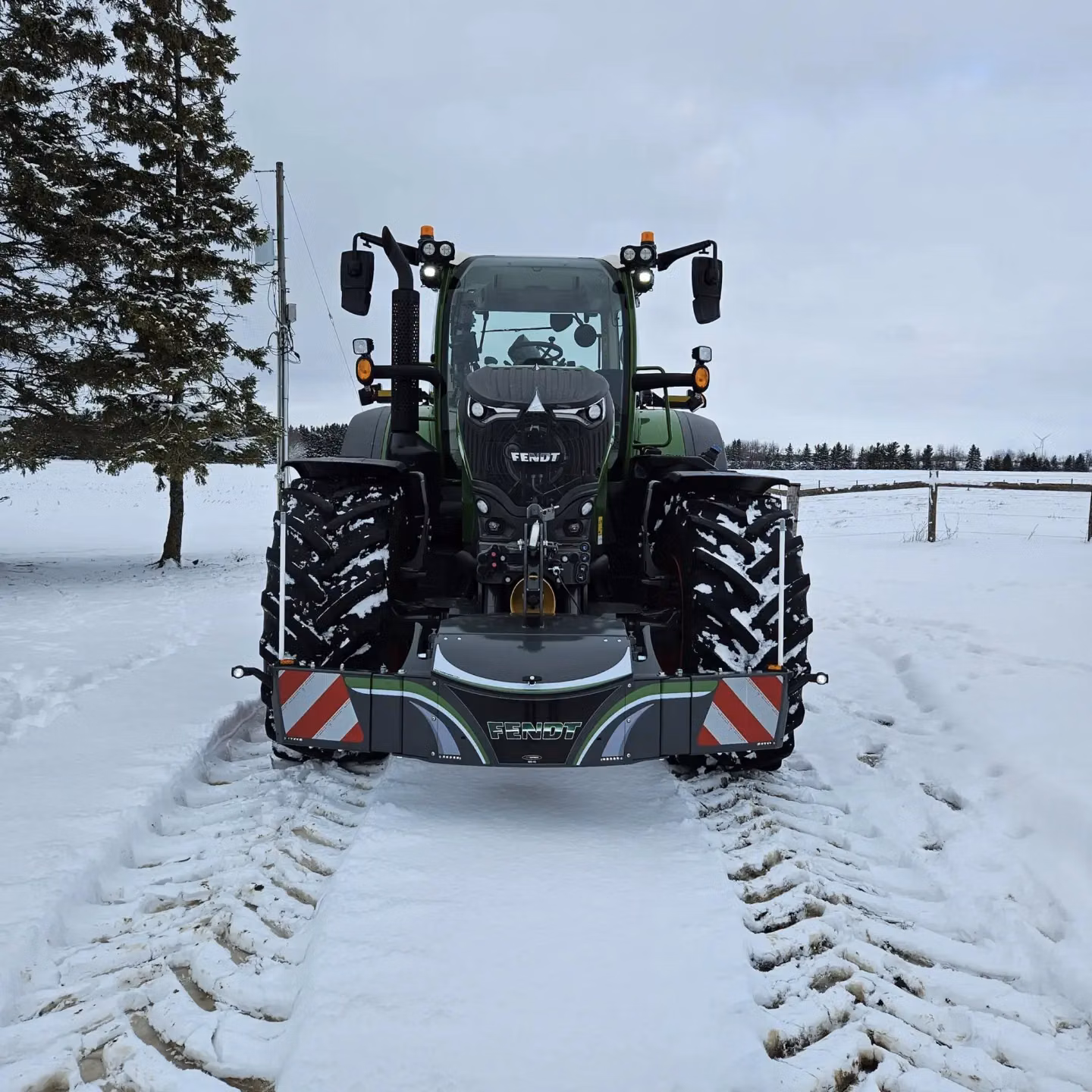 A green tractor with a front weight bumper featuring integrated lights and reflective safety panels on a snowy field