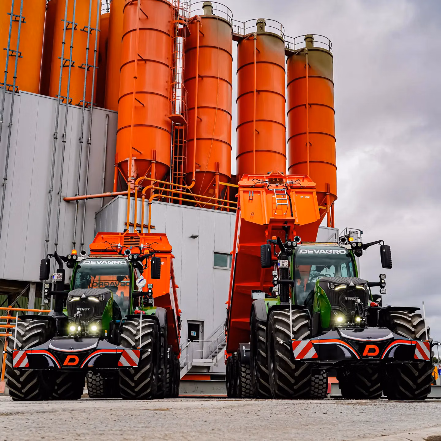 Two green tractors with large front weight bumpers and attached orange trailers parked by tall orange silos