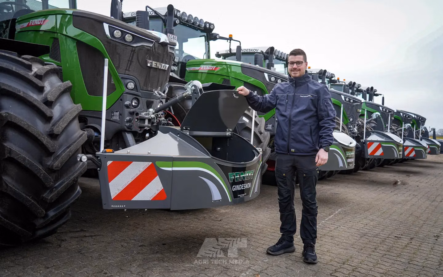 Man standing beside a green tractor fitted with a front weight bumper that has an open compartment and safety markings