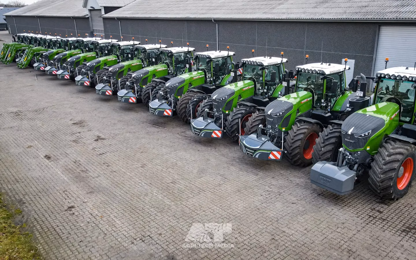 A row of green tractors parked outside, each fitted with large front weight bumpers featuring safety markings
