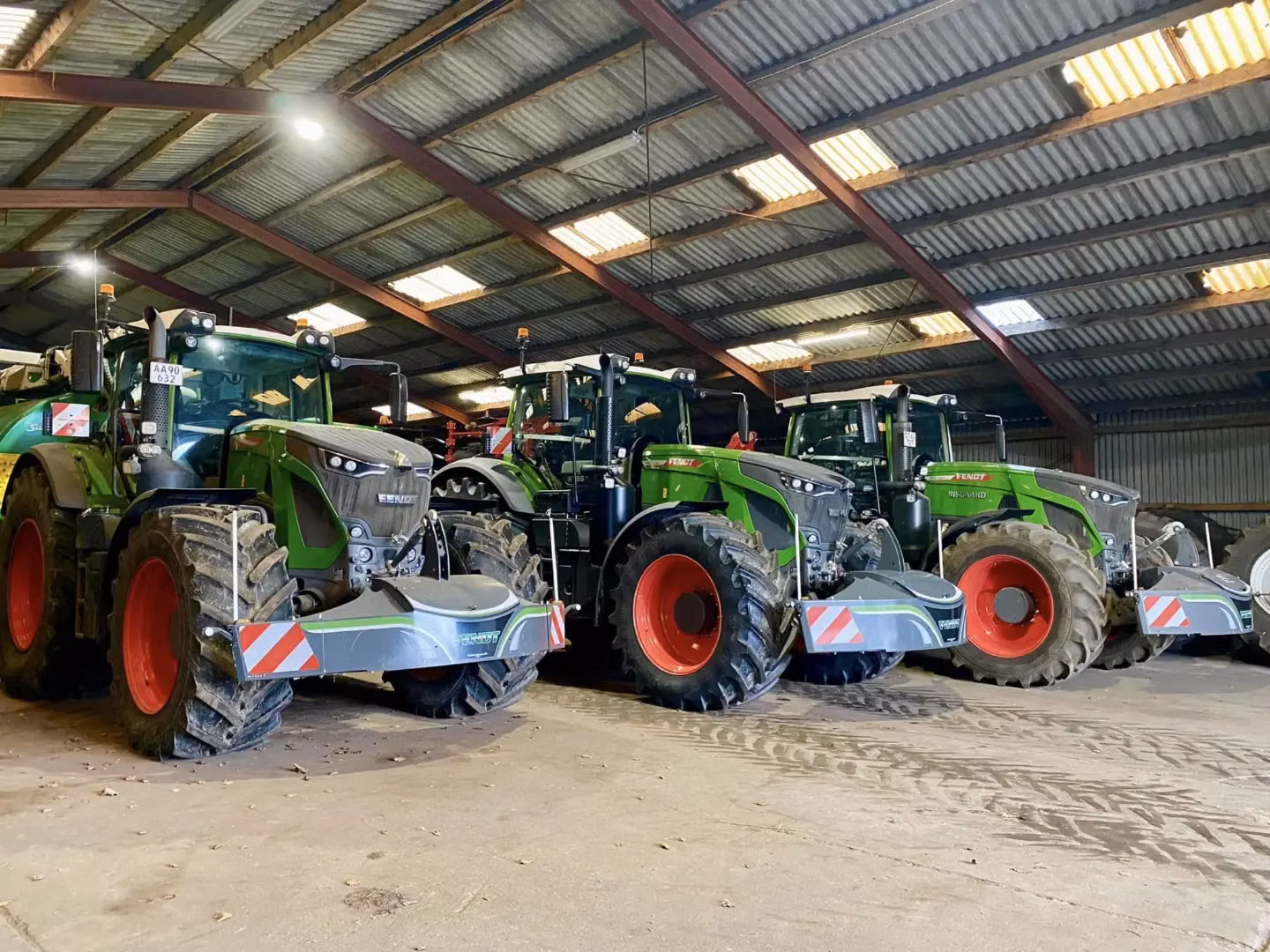 Four green tractors with front weight bumpers and red wheels parked inside a large shed