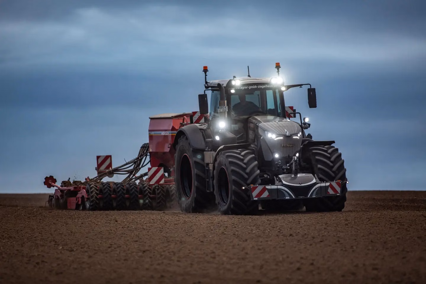 Large tractor with a front weight bumper and attached machinery driving over a plowed field at dusk