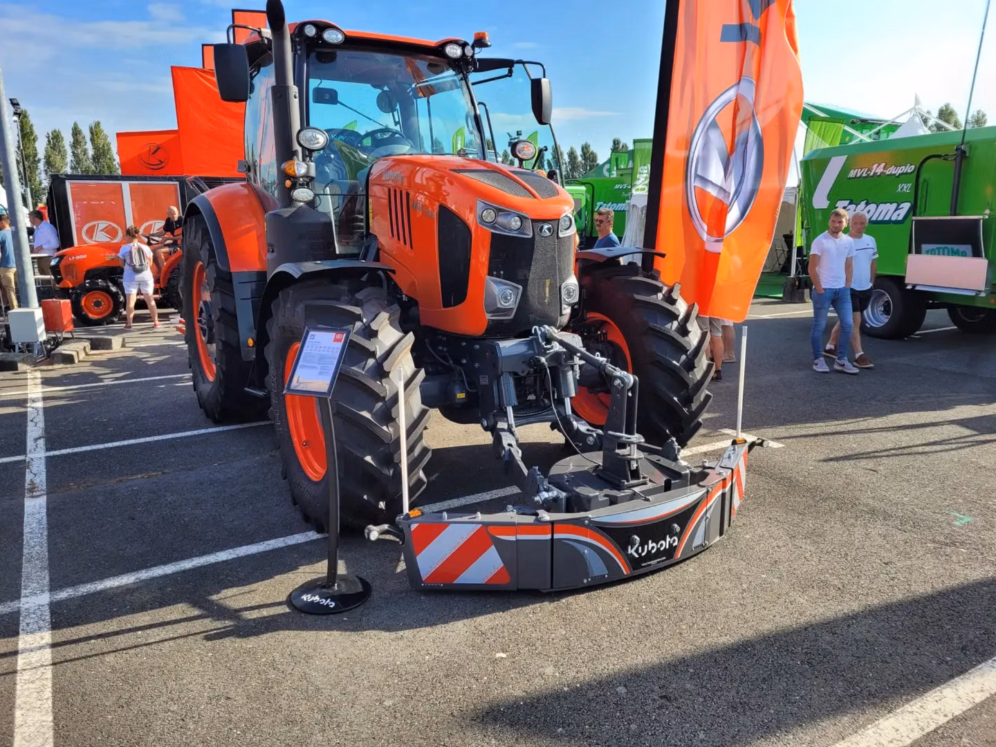 Large orange tractor with a front weight bumper attached, displayed outdoors with people and banners nearby
