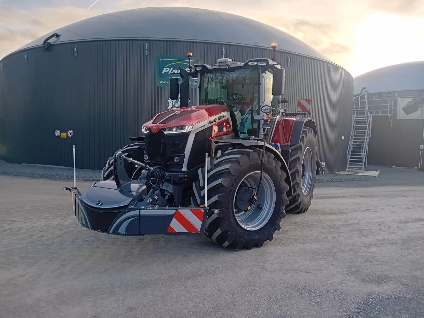 Red and black tractor with a front weight bumper attachment parked on gravel in front of large round metal buildings