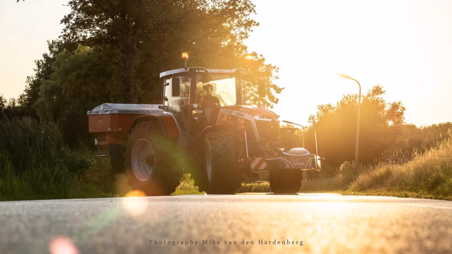 Tractor with a mounted front weight bumper driving on a rural road at sunset with orange warning lights on