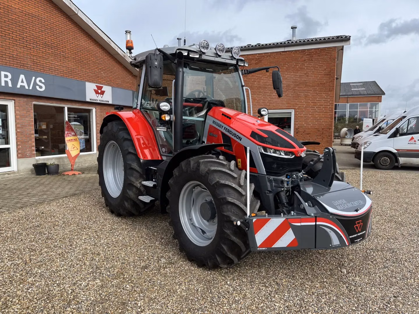 A red tractor with a large gray front weight bumper featuring red and white stripes is parked on gravel outside a brick