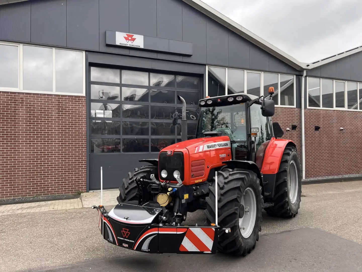 A red Massey Ferguson tractor with a black and red front weight bumper is parked outside a building with large windows